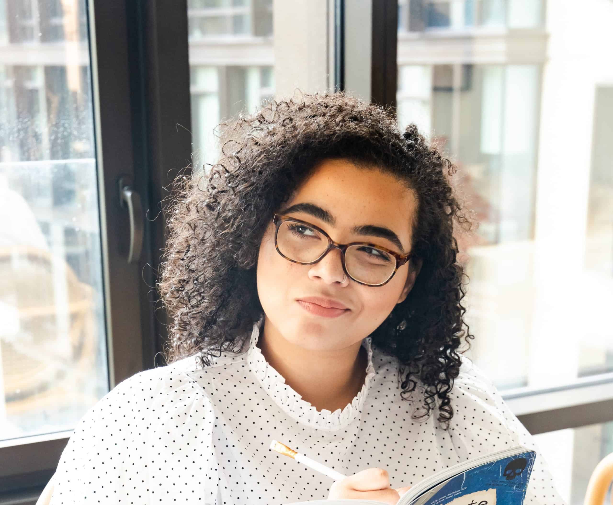 A woman with dark brown short curly hair, wearing tortoise shell glasses has a pen in her right hand on a notebook in front of her and a questioning look on her face as she journal about ditching diet culture and intuitive eating practices