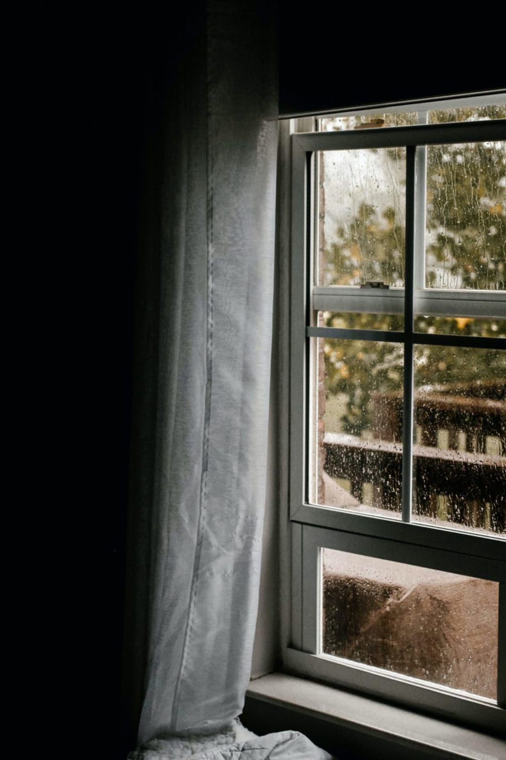 Dark room with light coming in through a glass pain window which has raindrops running down it. Looks out to trees and a deck barely visible and a grey rainy day. A white curtain hangs to the left of the window down past it's bottom.