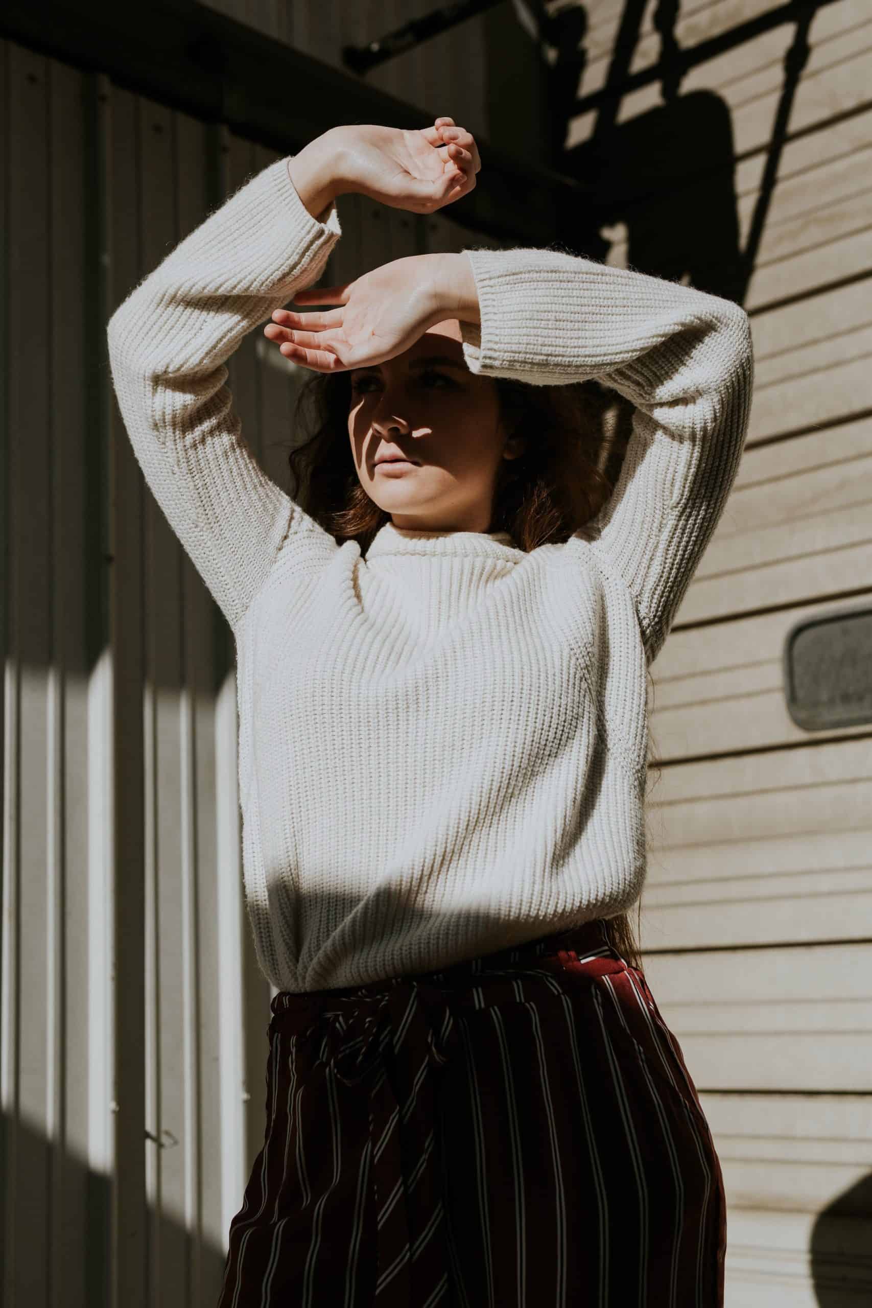 Woman in long sleeve white knitted top with arms lifted above her to block the sun she is facing. Shadows cast on her and around her.