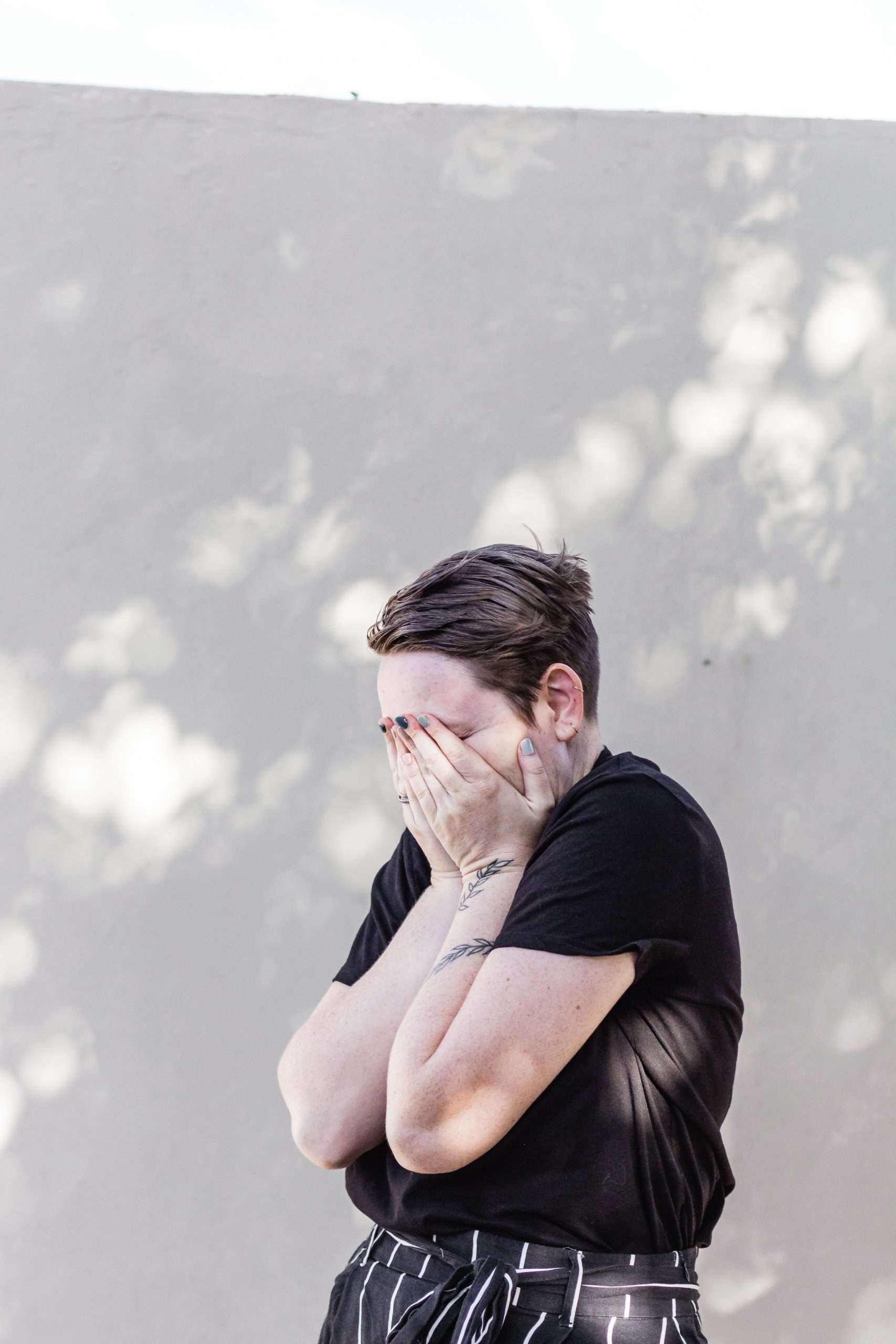 White person with short brown hair standing in front of while wall holding their face in their hands. They are wearing a black short sleeved shirt and black pants wiht white vertical stripes. There is a tattoo of a vine on left forearm and wrist. Their face is hidden in their hands.