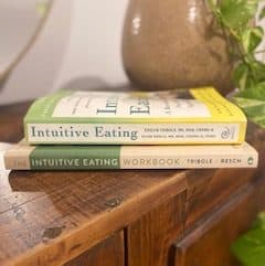 View of spines of stacked books. Intuitive Eating Book 4th edition stacked on top of the Intuitive Eating Workbook. Books sit on top of dark wood cabinet. Green crawling potted plan in grey pot behind. 