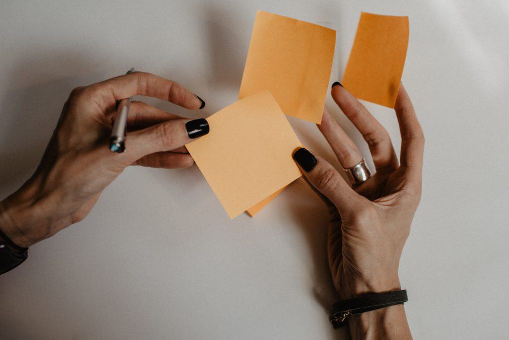 slender older womans hands with black nail polish holding three orange sticky notes between fingers and a pen in the left hand. Black bracelet on right wrist. Contemplating writing down a positive body affirmation.