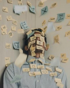 Man standing in corner covered by small and large blue sticky notes and yellow sticky notes with writing and doodles on them in black sharpee