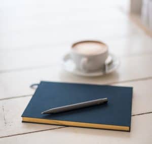 Navy blue notebook with black pen sitting on top of it sitting on a white wood panel table. Latte in white tea cup resting in white saucer sits behind it. 