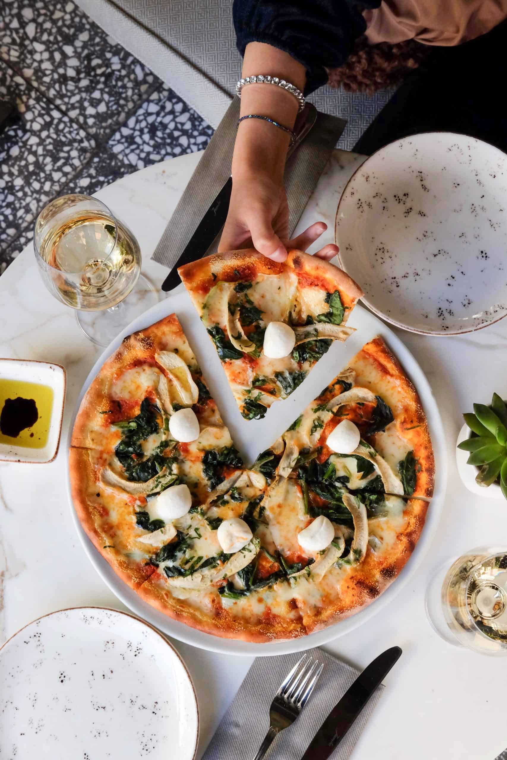 large pizza on a table with a woman who has made peace with pizza and has permission to eat all foods taking a slice of pizza off the plate. Pizza surrounded by two empty white plates with black speckles, two glasses of white wine, some silverware and dish of olive oil and balsamic vinegar.