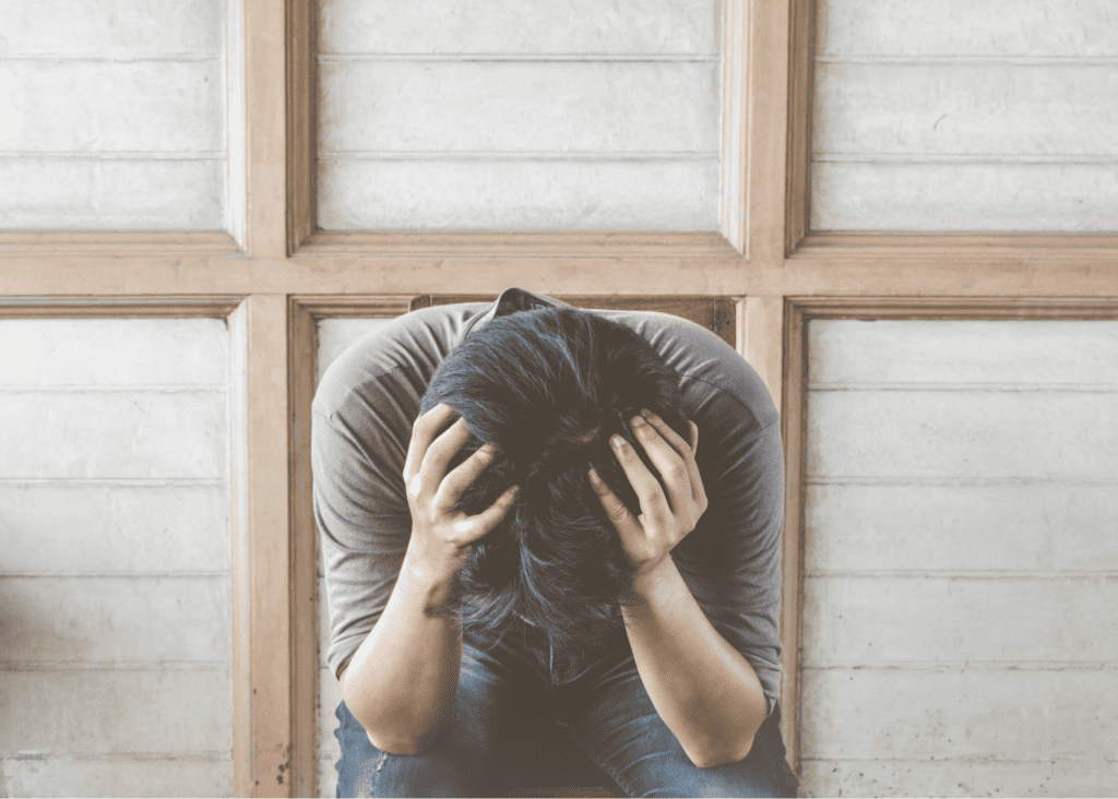 person with black hair, gray mid length t-shirt and blue jeans on. Their elbows on knees, hunched over with head in hads