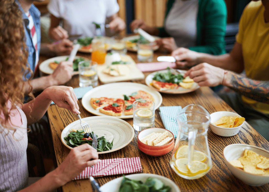 wooden table with variety of food and drinks spread out. Several sets of hands visible of people seated and eating at the table. 