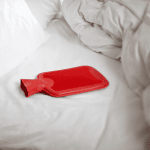 red hot water bottle resting in white bed sheets