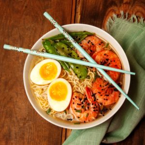 Bowl of yellow ramen noodles topped with orange shrimp with black sesame seeds on 1/3, a soft boiled egg cut in half with yolk facing up on 1/3 and edemame beans on the other 1/3. Sitting on top of green cloth napkin and hard wood table