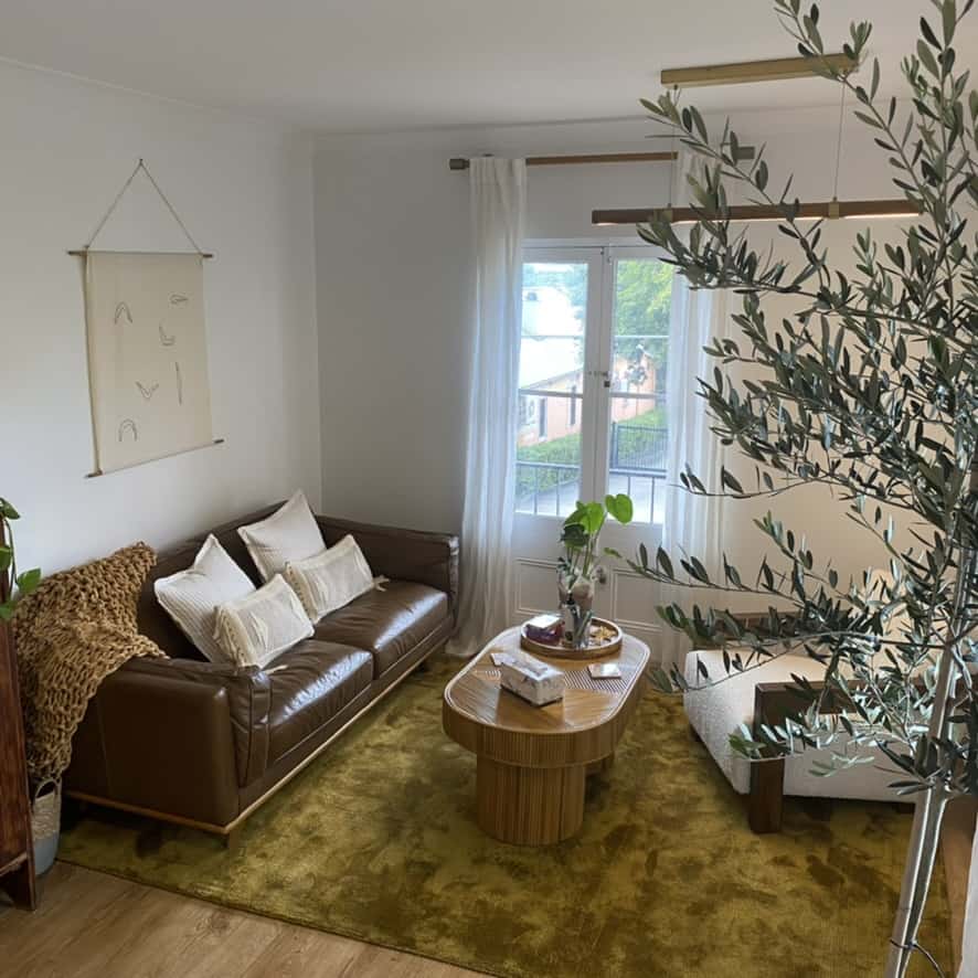 ACD55AD4-0284-498B-9638-B70C1DC3F5A4_1_105_c Image of inside the non Diet and weight inclusive Dietitian Clinic Wander and Nourish Nutrition. Golden rod rug, brown leather couch, olive tree, bamboo coffee table and sheer curtains with sunlight streaming through the window.