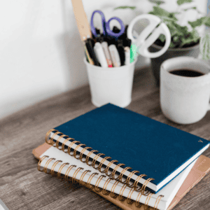 navy spiral bound notebook on top of a white one on top of a brown folder sitting on wooden table. Cup of pens sits behind and cup of coffee.