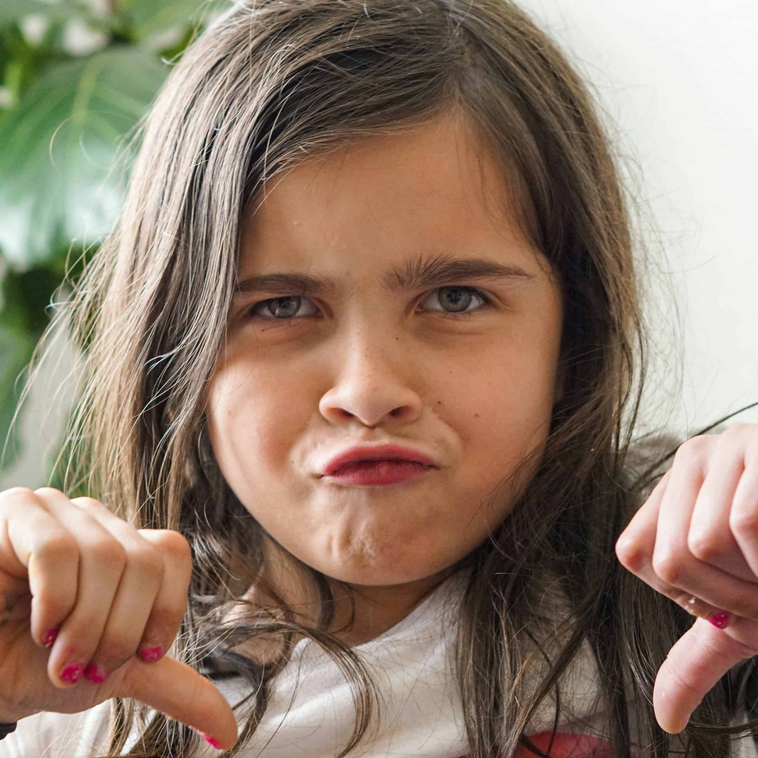 A little caucasian girl with brunette hair, around age 8, making a frowning face and hands in front of her with thumbs pointed down